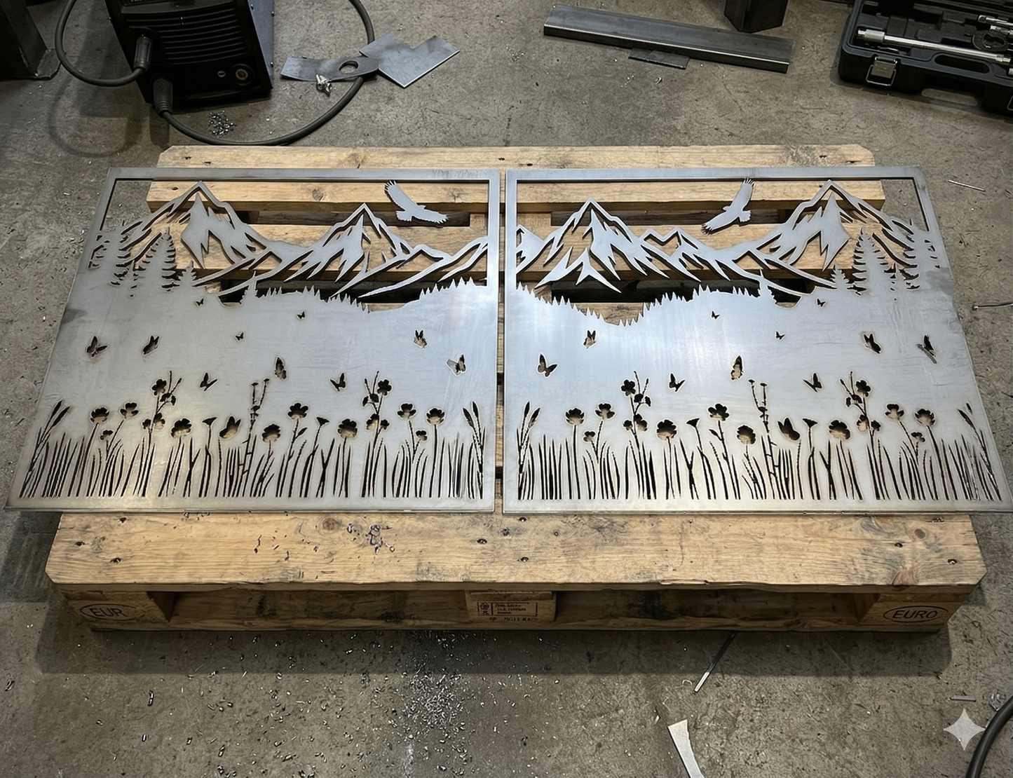  Real-life photo of raw metal panels with a laser-cut mountain and eagle motif, resting on a pallet in a metal fabrication workshop.