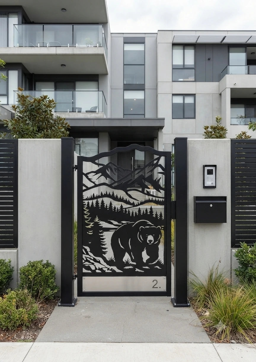 A modern black metal residential gate with a laser-cut bear and mountain landscape design, installed on a property boundary in front of a contemporary apartment building.